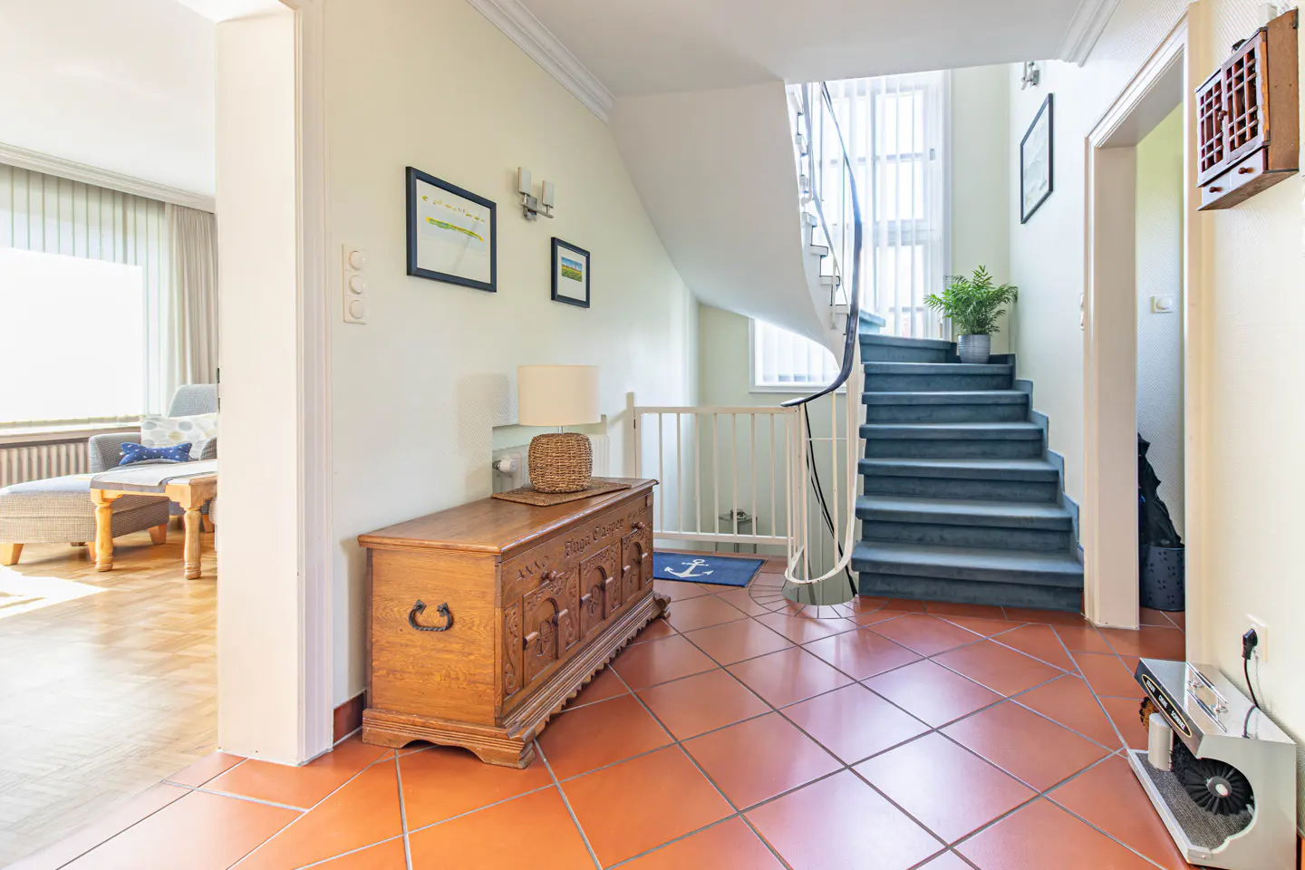 Entryway with terracotta tile floor, wooden chest, and blue carpeted stairs with a white spiral banister. A living room is visible through an opening.