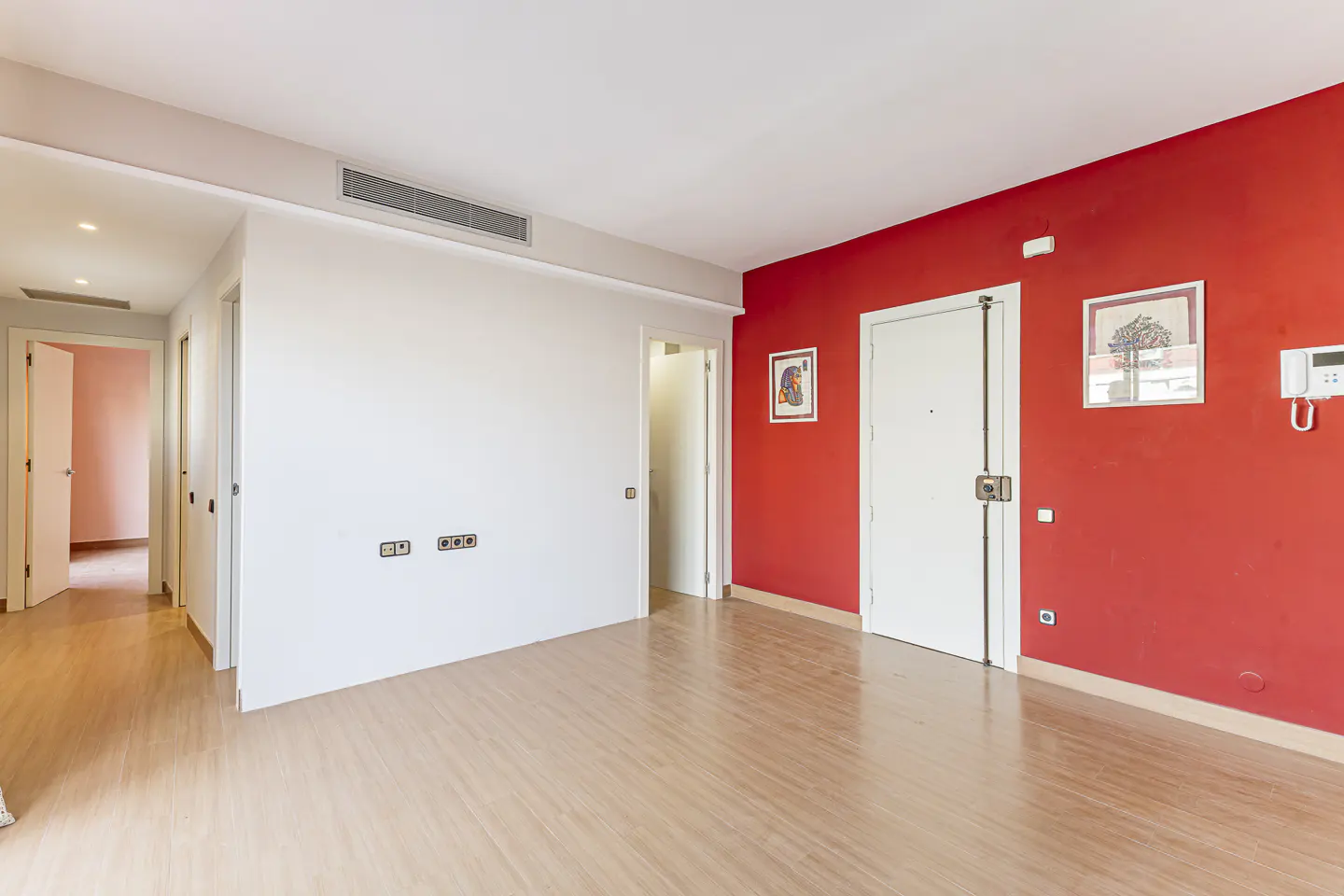 Bright, empty room with wood floors, white and red walls. Hallway visible on the left. Two framed pictures on the red wall.