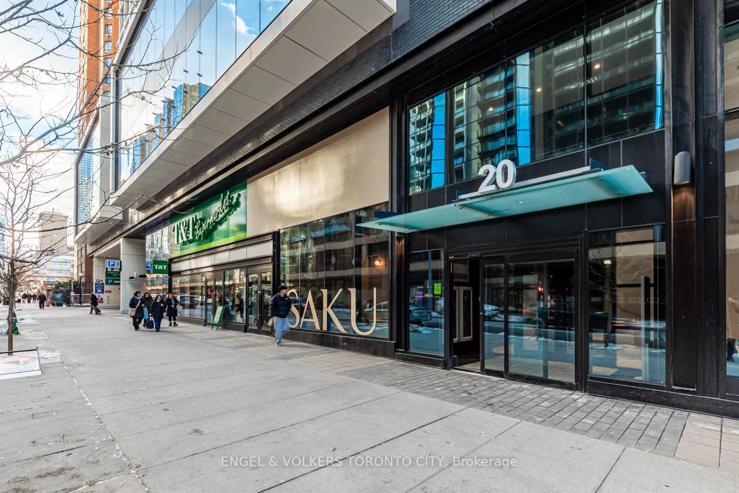 Street-level view of a modern building entrance at 20 with T&T Supermarket and Saku signage, people walking on the sidewalk.