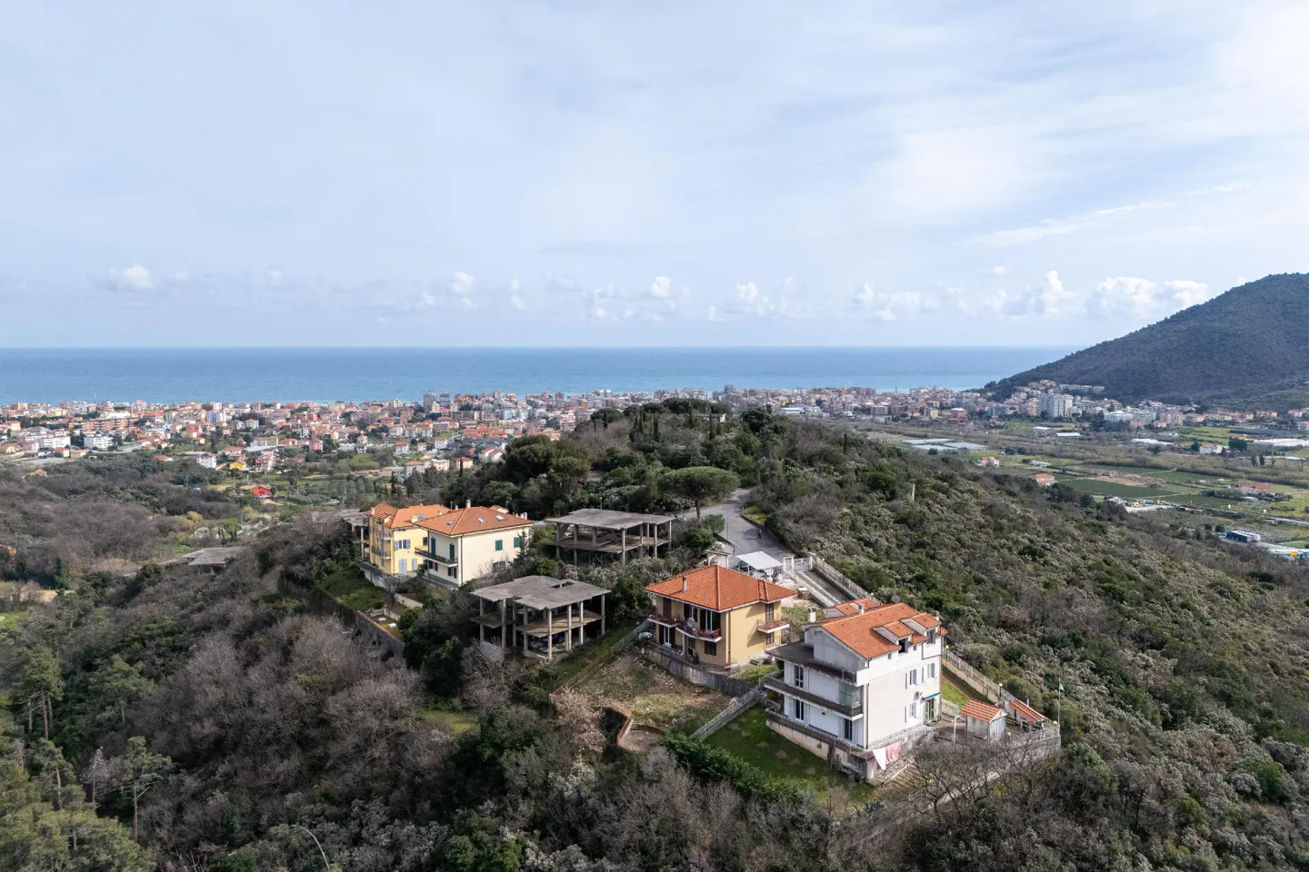 Aerial view of houses on a wooded hillside overlooking a coastal town and the sea under a cloudy sky.