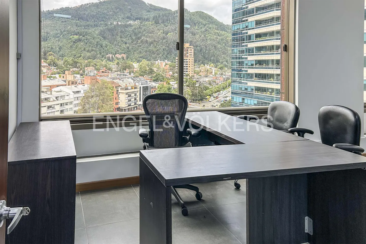 Office interior with a large desk, chairs, and a view of a city with mountains in the background.