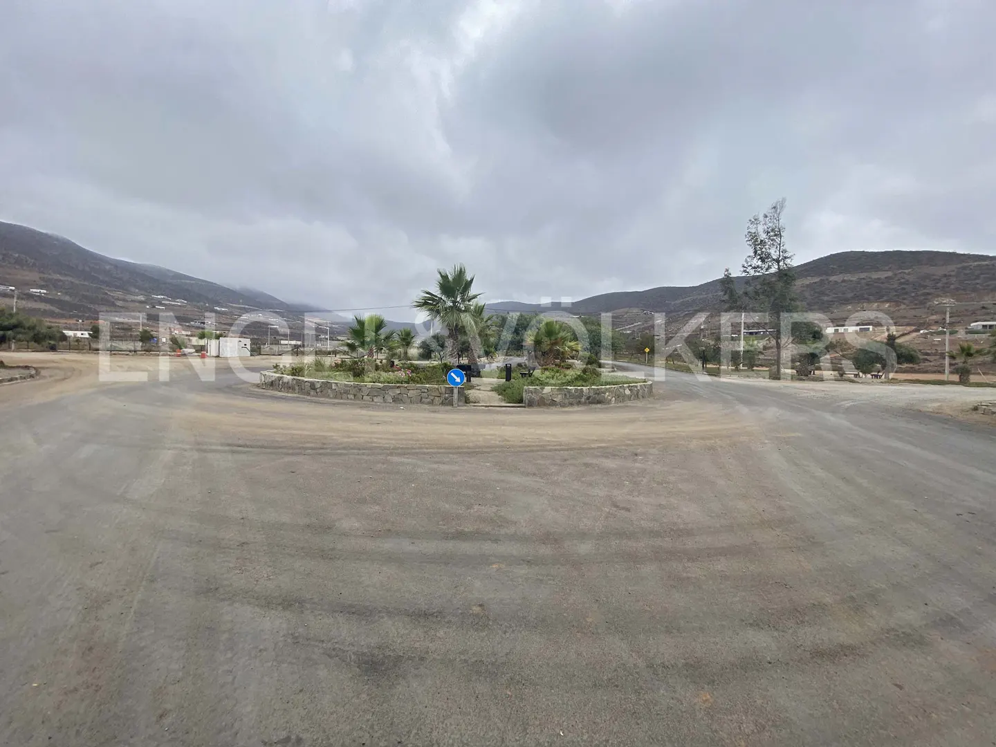 A roundabout with palm trees and a blue directional sign, surrounded by roads and hills under a cloudy sky.