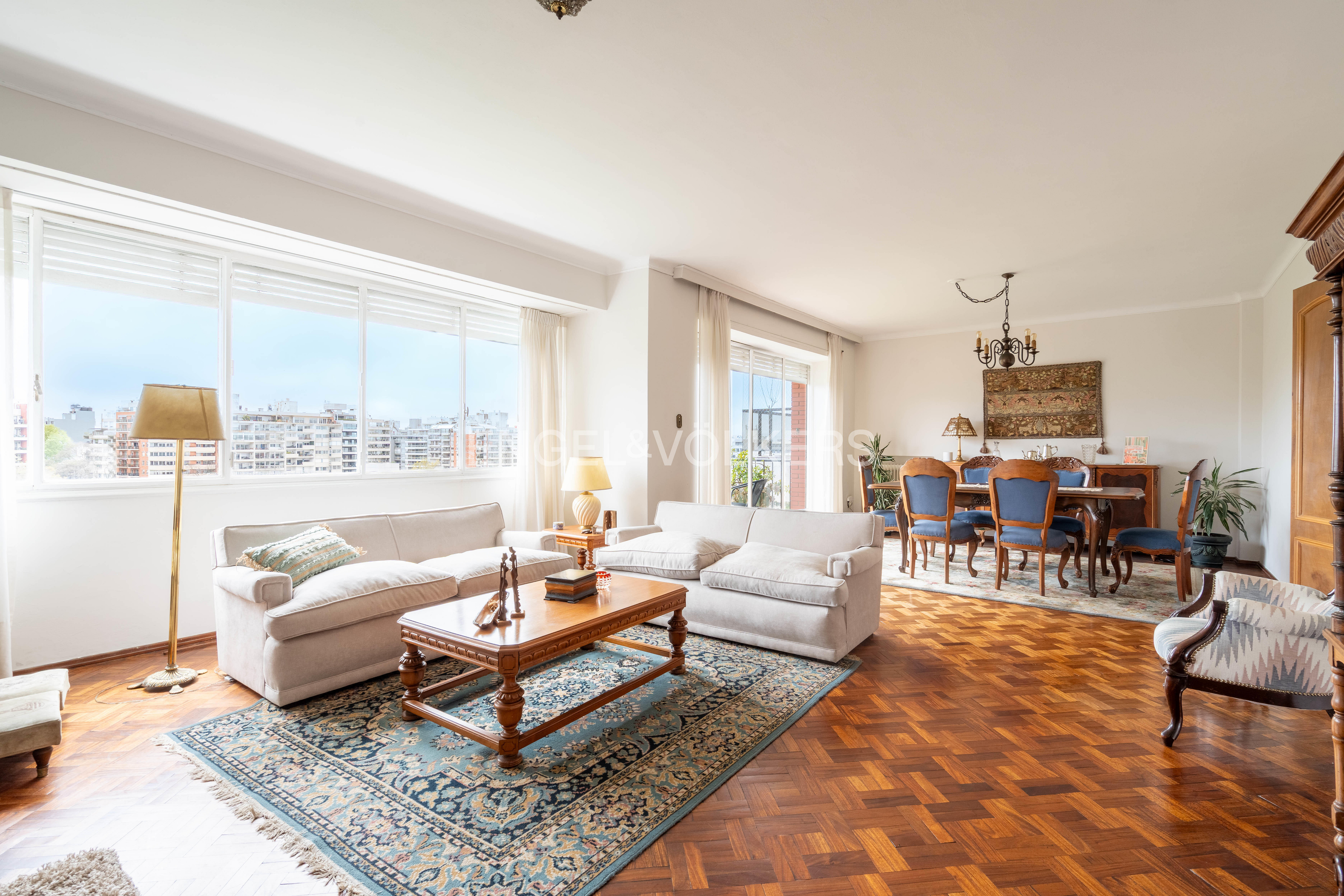 Bright living room with white sofas, wooden coffee table, patterned rug, and a dining area with blue chairs on wood floors. [AI-generated text]