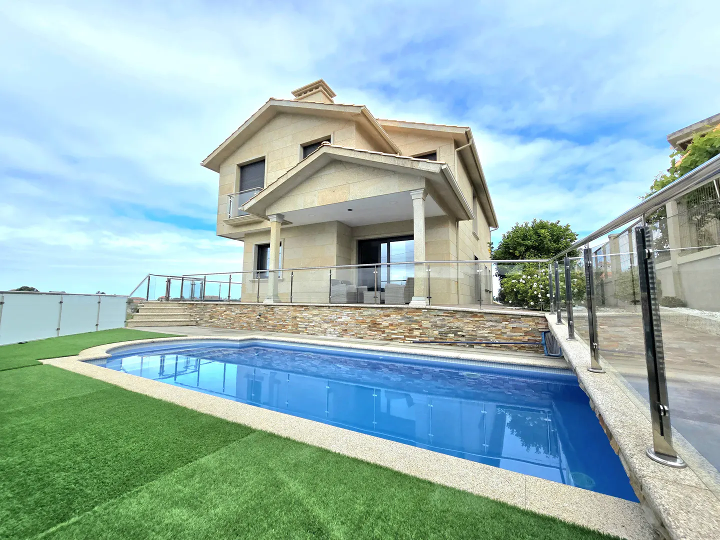 A two-story tan house with a blue swimming pool and green artificial grass.
