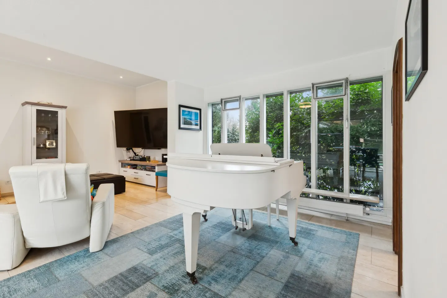Bright living room with a white grand piano on a blue rug, a white recliner, and a wall of windows.
