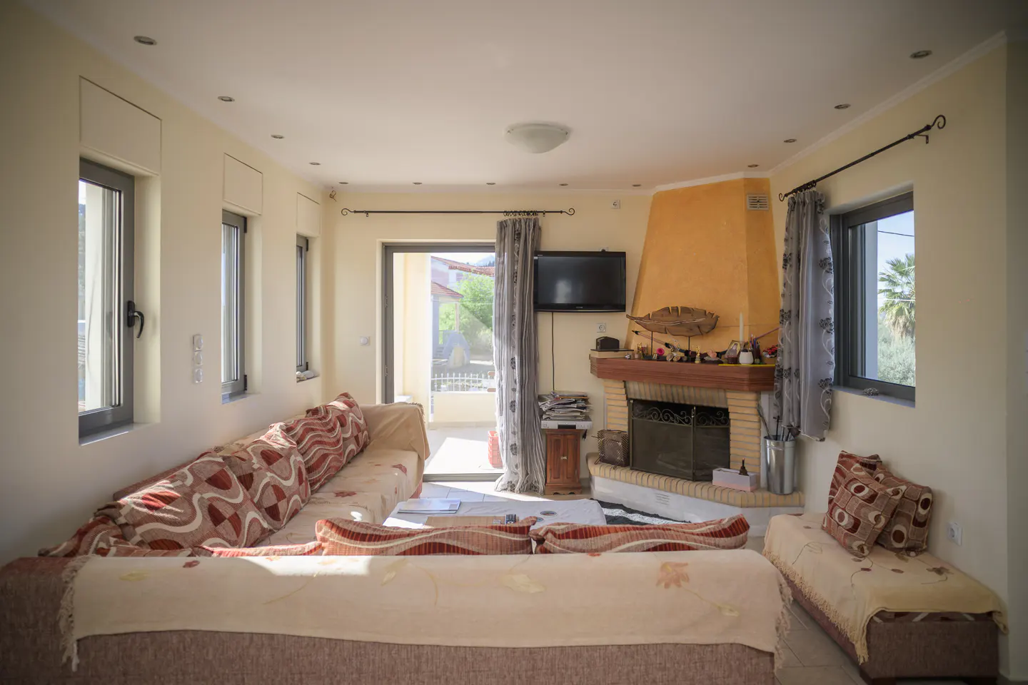 Living room with beige walls, a brick fireplace, and a tan sofa with patterned pillows. Windows and a sliding glass door let in natural light.