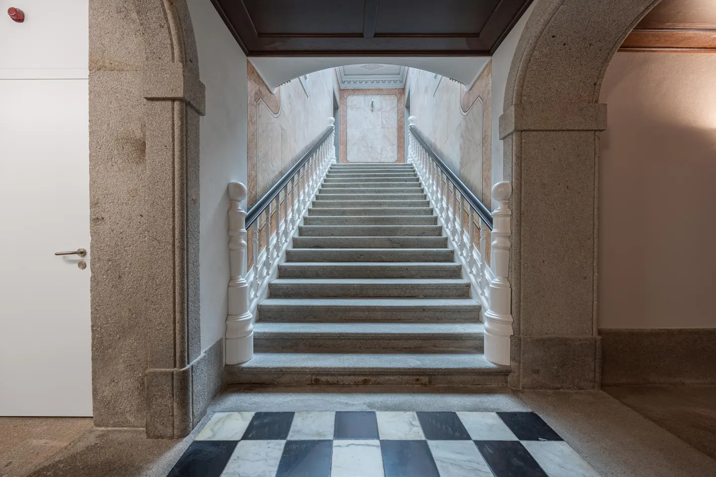 Grand staircase with stone steps, white balusters, and black handrail. Black and white checkered marble floor. Arched stone doorway.