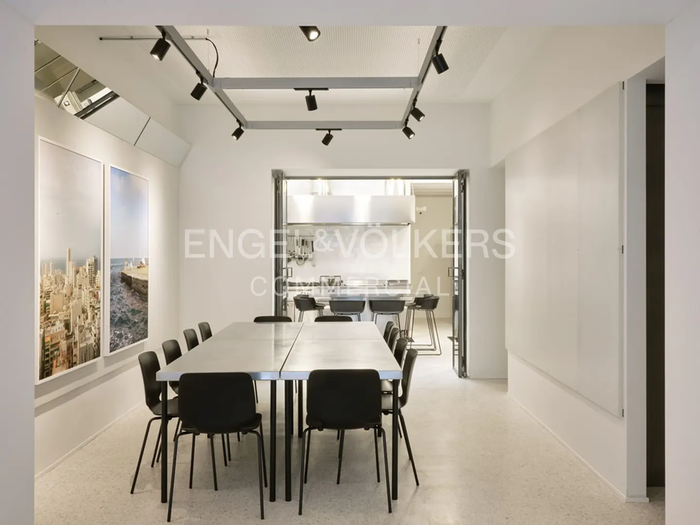 Bright, modern office space with a long table, black chairs, and track lighting. A kitchen is visible through an open doorway. Two framed pictures hang on the wall.