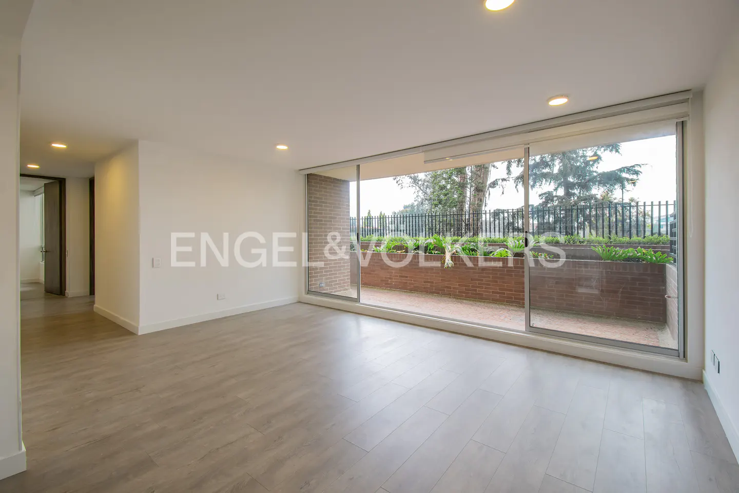 Empty living room with gray wood floors and white walls. Large sliding glass doors open to a brick patio with greenery and a black metal fence.