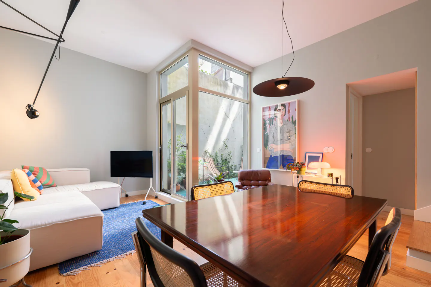 A modern living room with a wood dining table, white sofa, blue rug, and a large window overlooking a small courtyard.