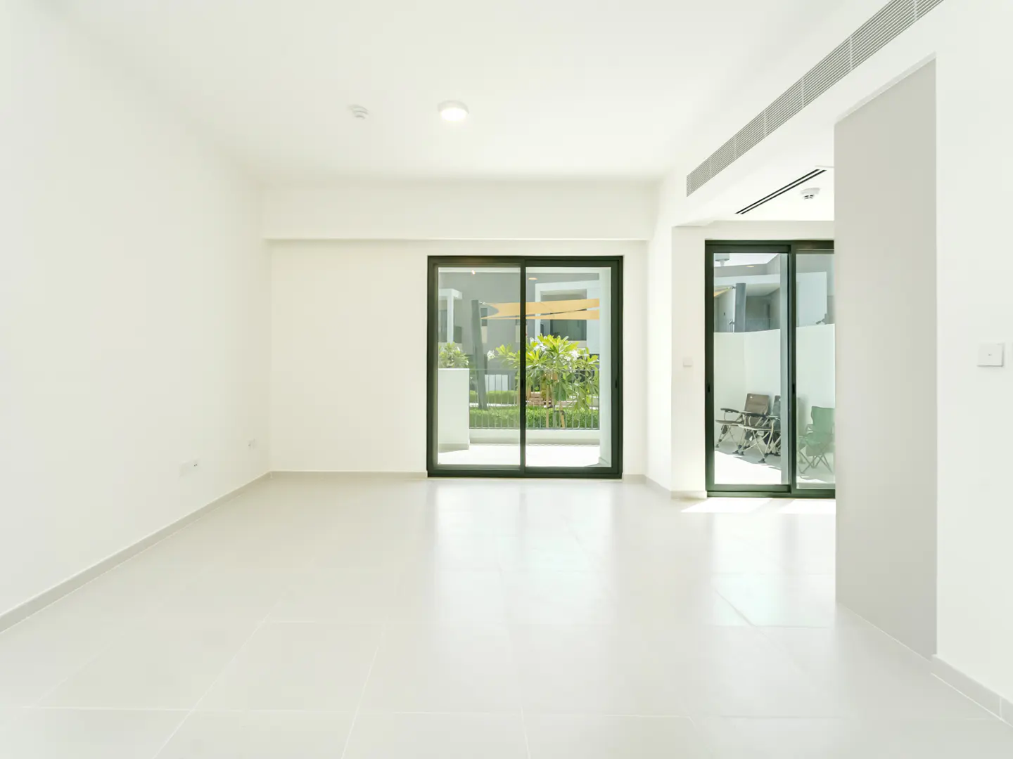 Bright, empty room with white walls and tile floor. Sliding glass doors lead to a patio with greenery and outdoor furniture.
