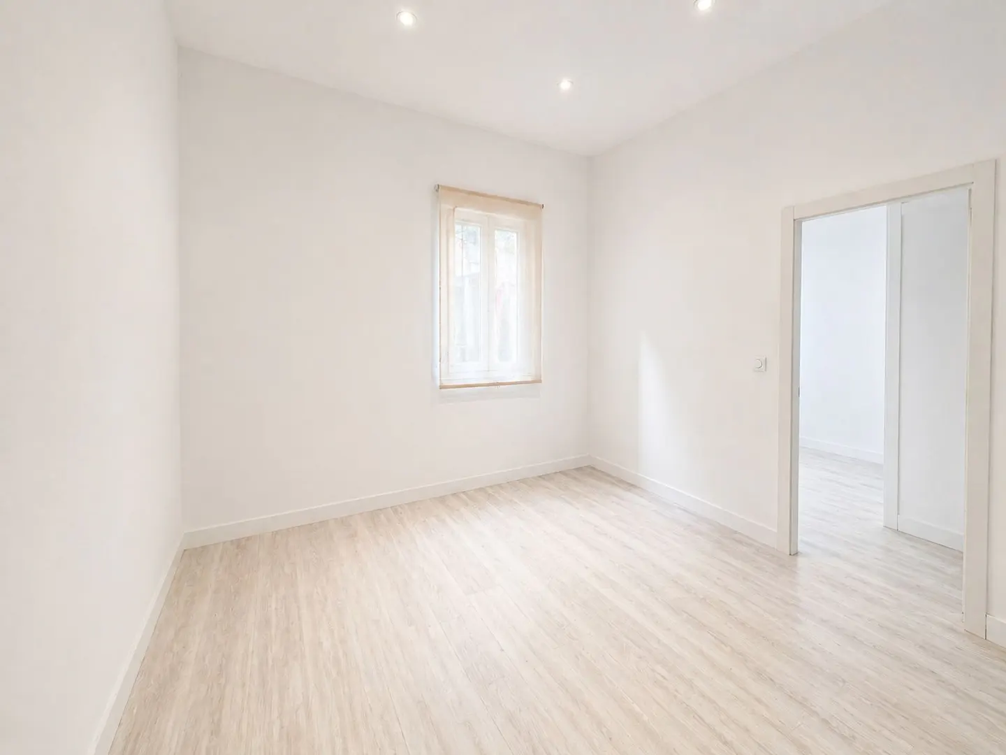 Bright, empty room with white walls, light wood floors, and a window with sheer curtains. A doorway leads to another room.