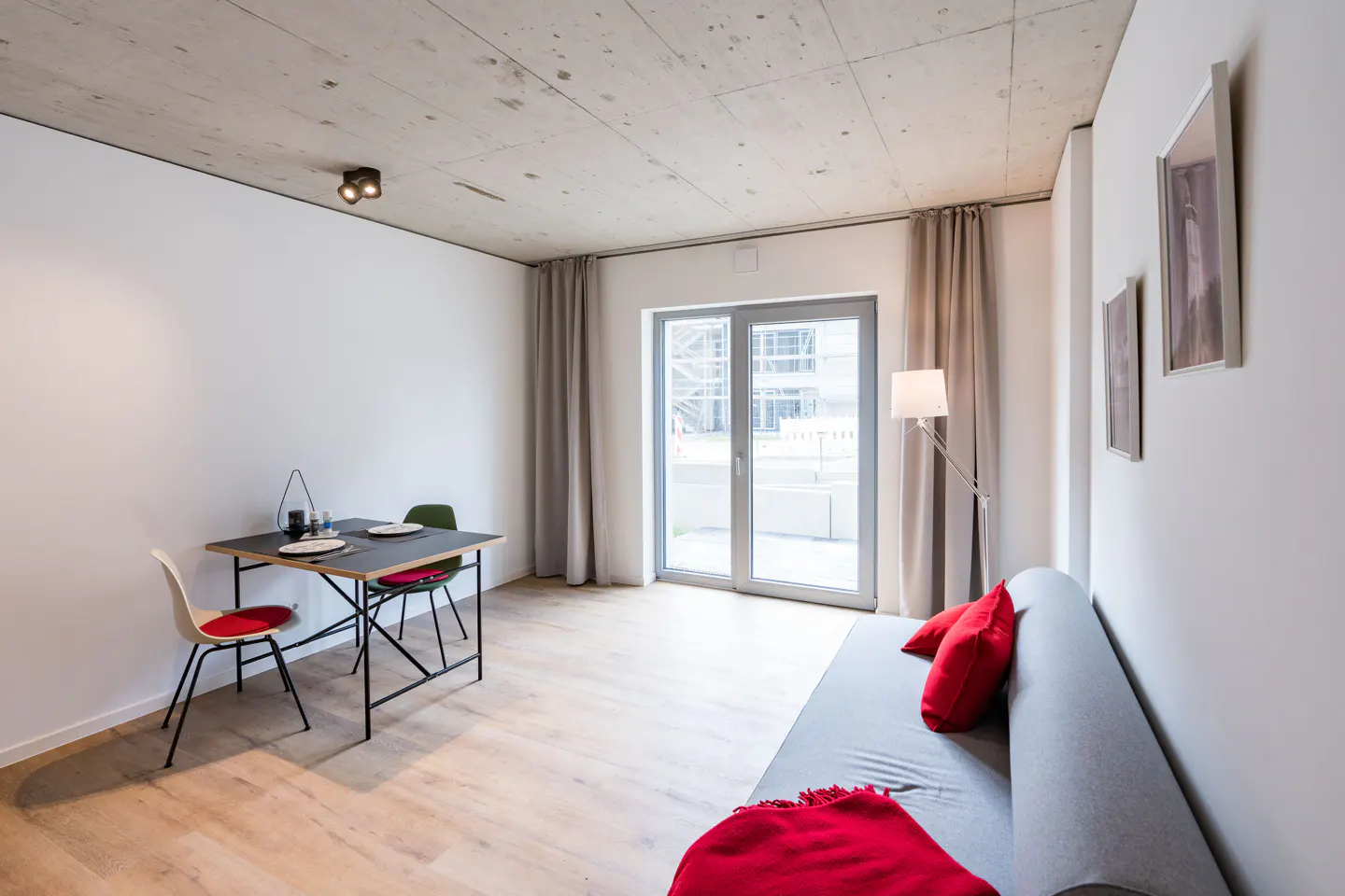 A modern apartment interior with a dining table set for two, a gray sofa with red pillows, and a glass door to a balcony.