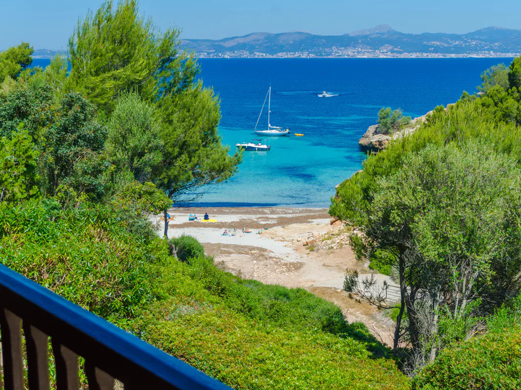 View of a turquoise Mediterranean cove framed by lush green trees, with sailboats anchored in the water and a sandy beach.