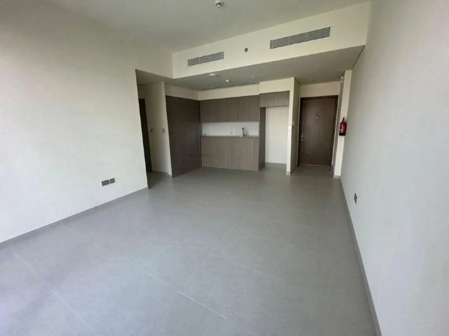 A bright, empty apartment with gray tile floors, white walls, and a small kitchen area with brown cabinets. A brown door and a red fire extinguisher are visible.
