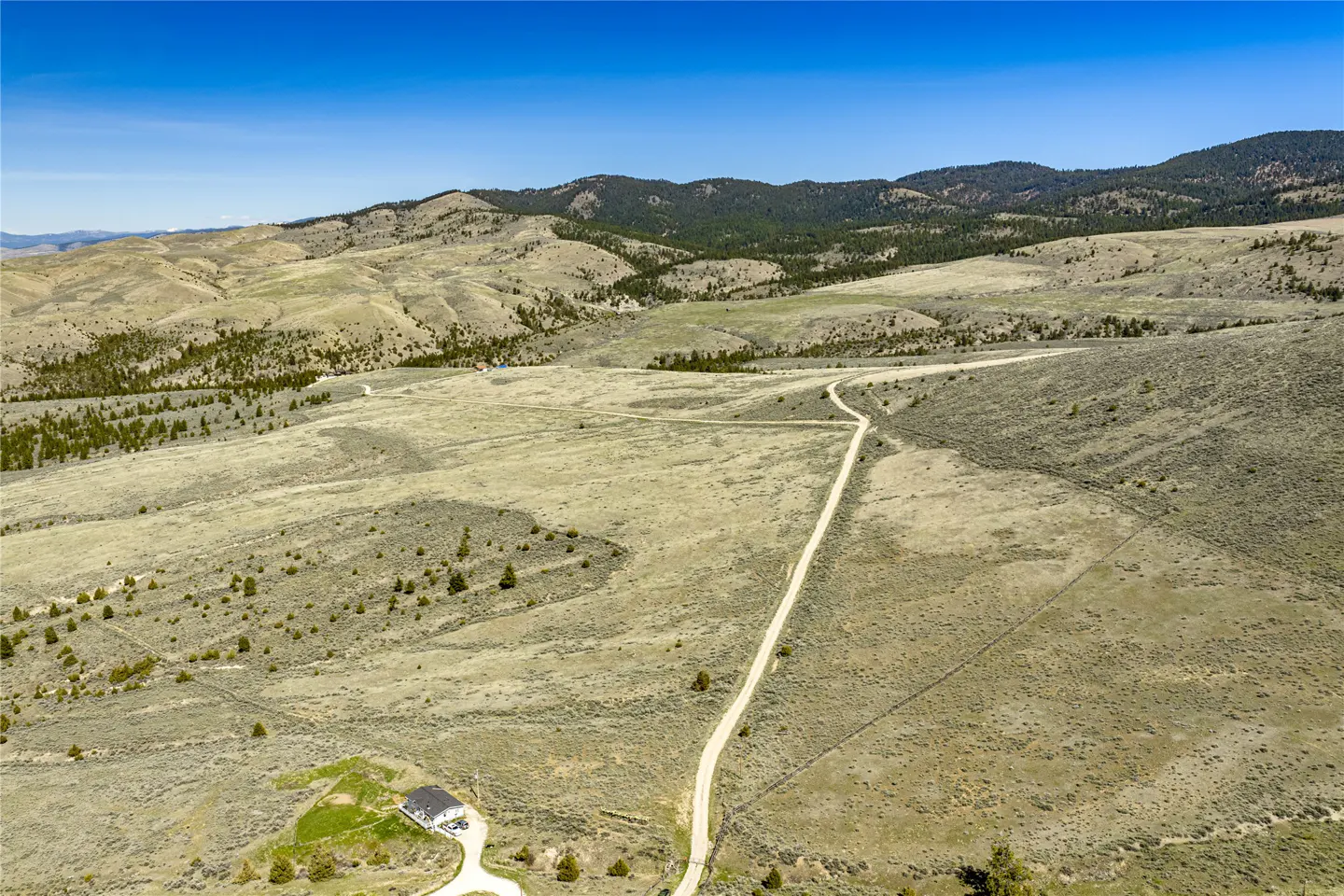 Aerial view of a house with a long dirt driveway in a grassy, hilly landscape under a blue sky.