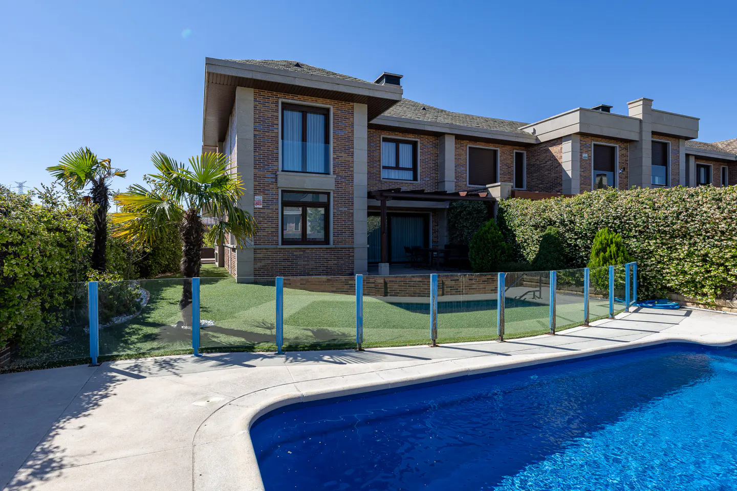 A two-story brick house with a backyard pool, glass fence, artificial turf, and palm trees under a clear blue sky.