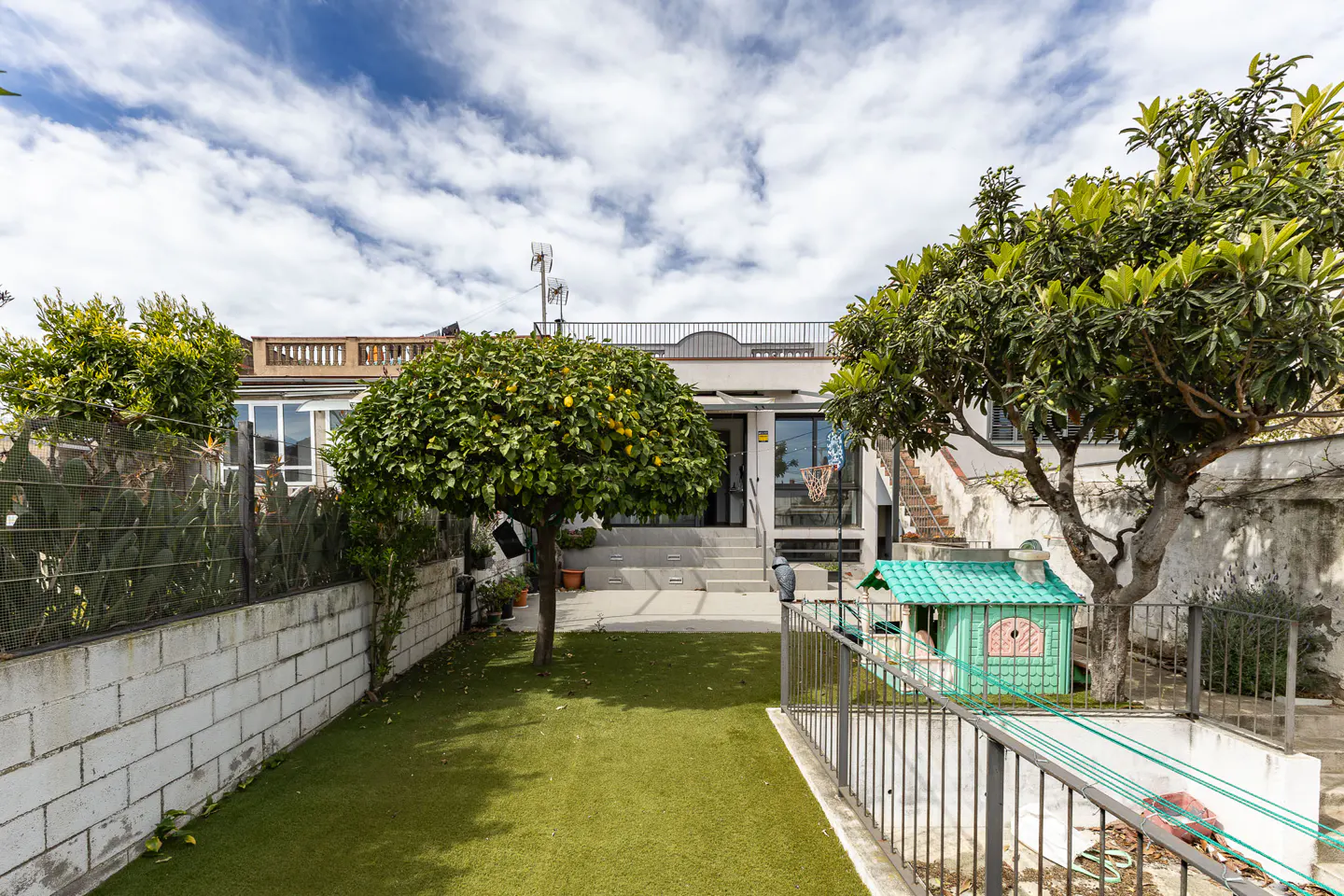A house with a green lawn, trees, and a playhouse. The house is white with a rooftop terrace. The sky is blue with white clouds.