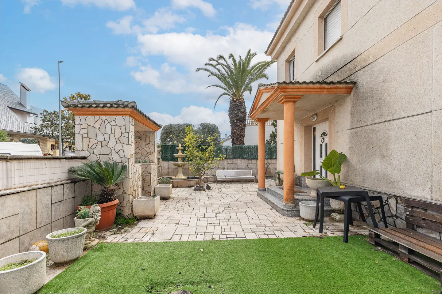 A beige two-story house with a stone patio, palm tree, fountain, and green lawn under a blue sky.