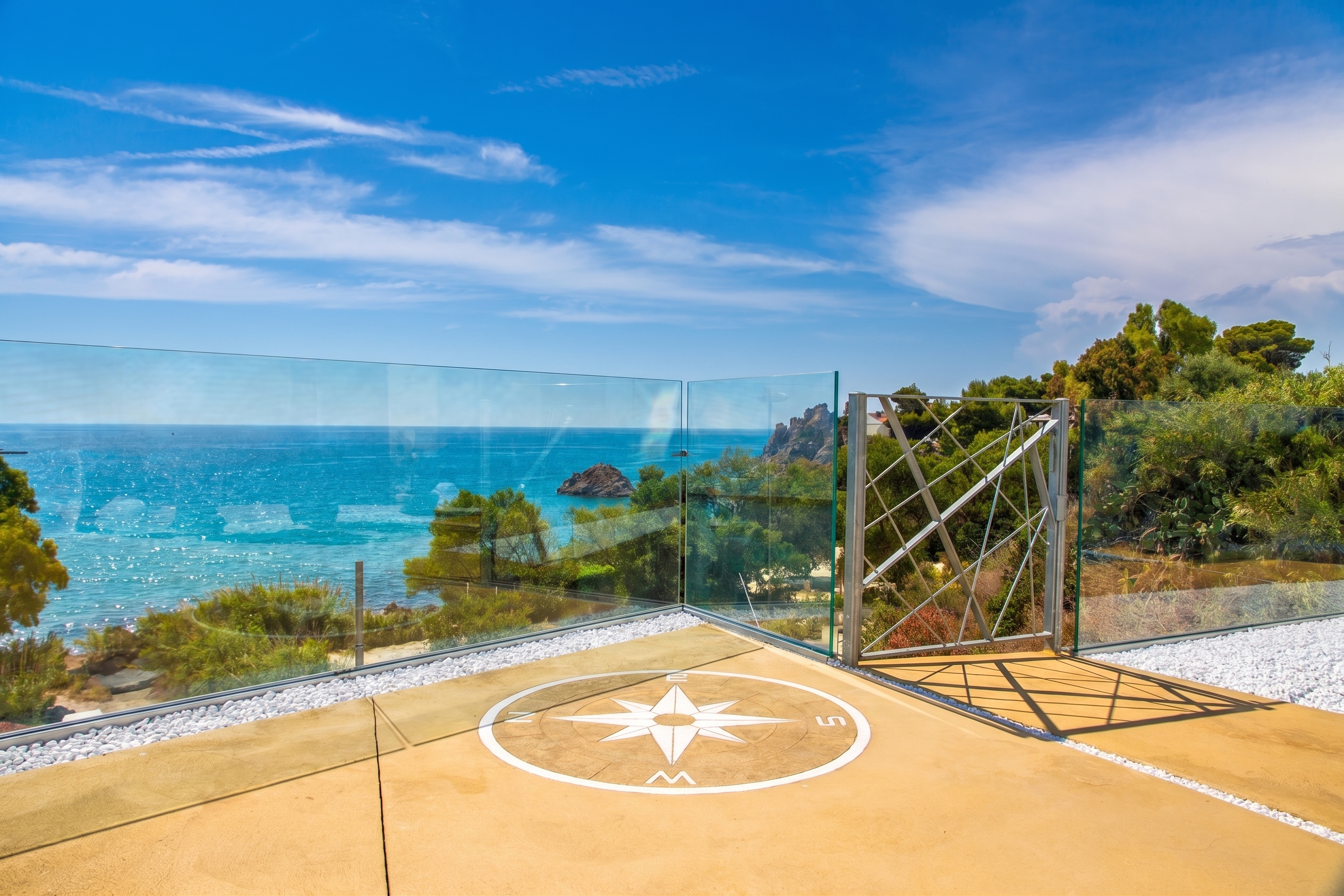 Terrace with a compass rose inlay, glass railing, and a metal gate overlooking the blue sea under a sunny sky.