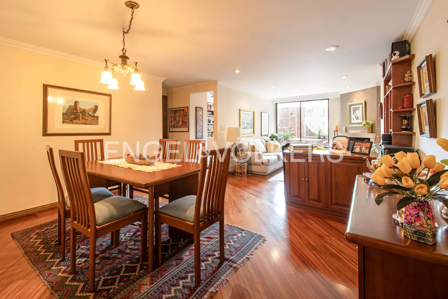 Dining area with wooden table and chairs on a patterned rug, opening into a living room with hardwood floors and fireplace.