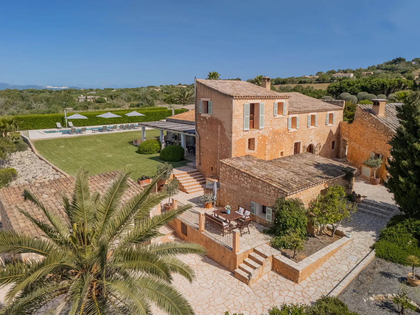 Aerial view of a large, orange stone house with a tile roof, a pool, and a green lawn on a sunny day.