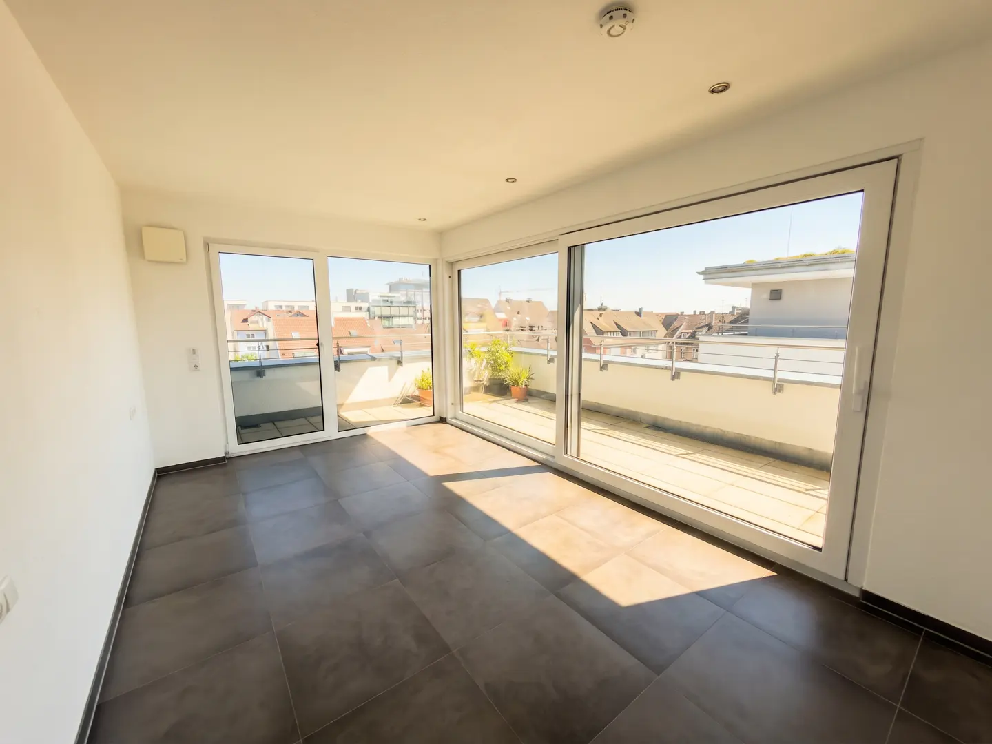 Bright, empty room with dark tile floors and white walls. Large sliding glass doors open to a sunny balcony with city views.