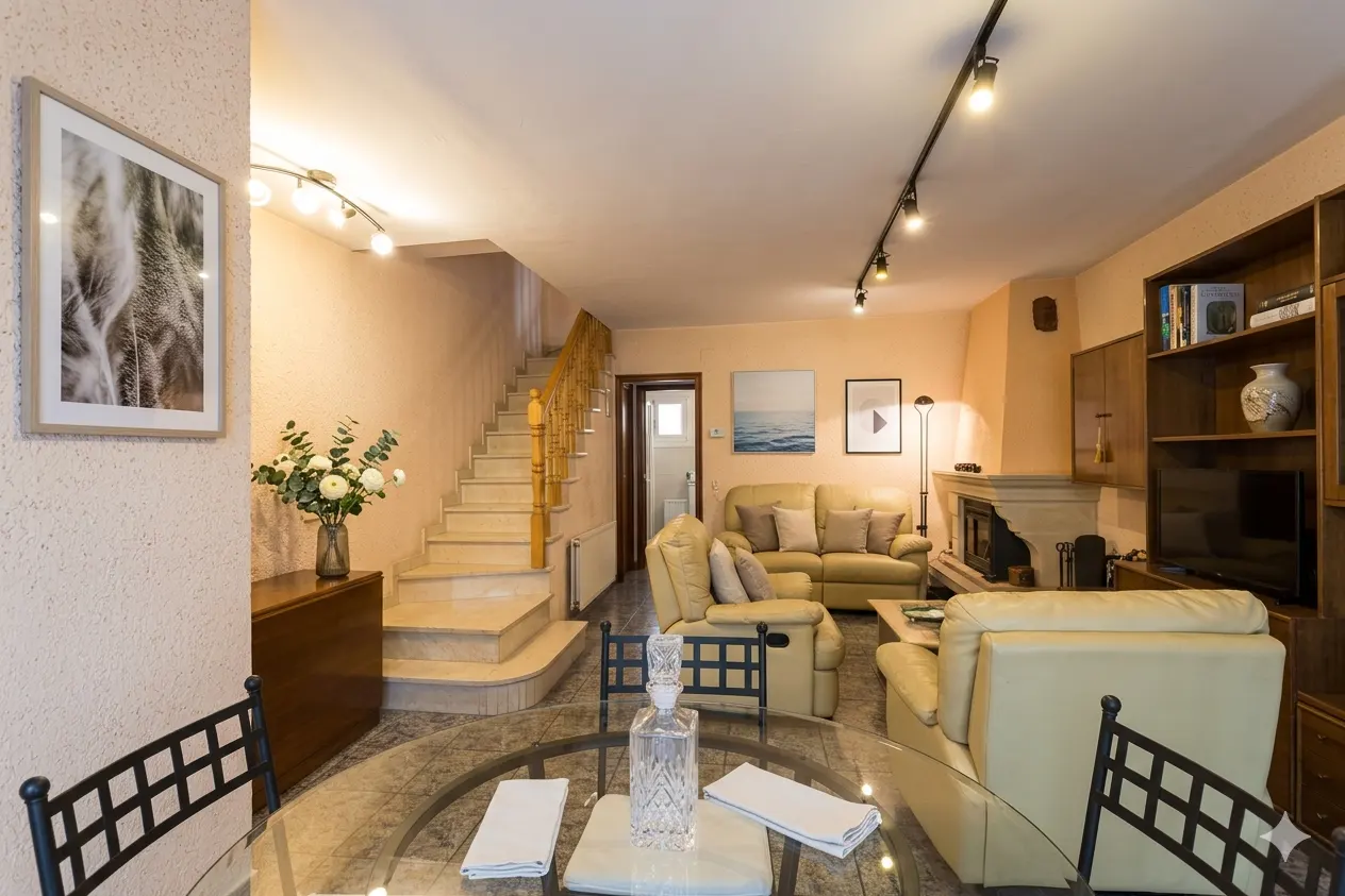 Living room with beige walls, a staircase, and a glass table with white napkins. A cream-colored sofa and fireplace are in the background.