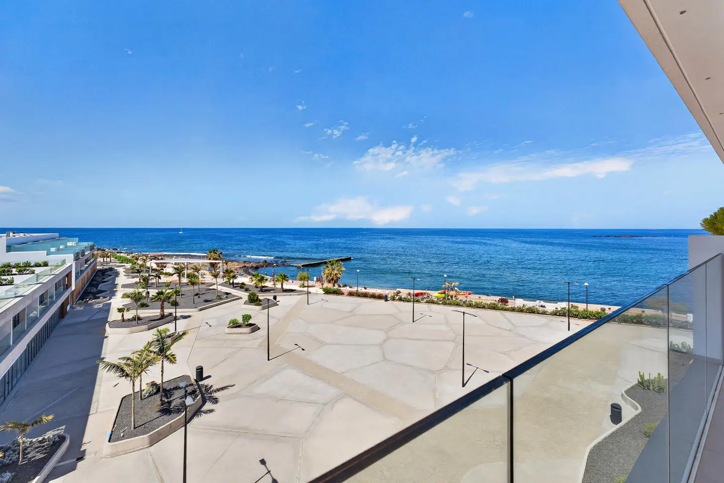 View from a balcony overlooking a plaza with palm trees, the ocean, and a blue sky.