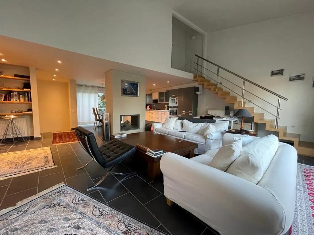 A bright, modern living room with white sofas, a black chair, and a wooden staircase.