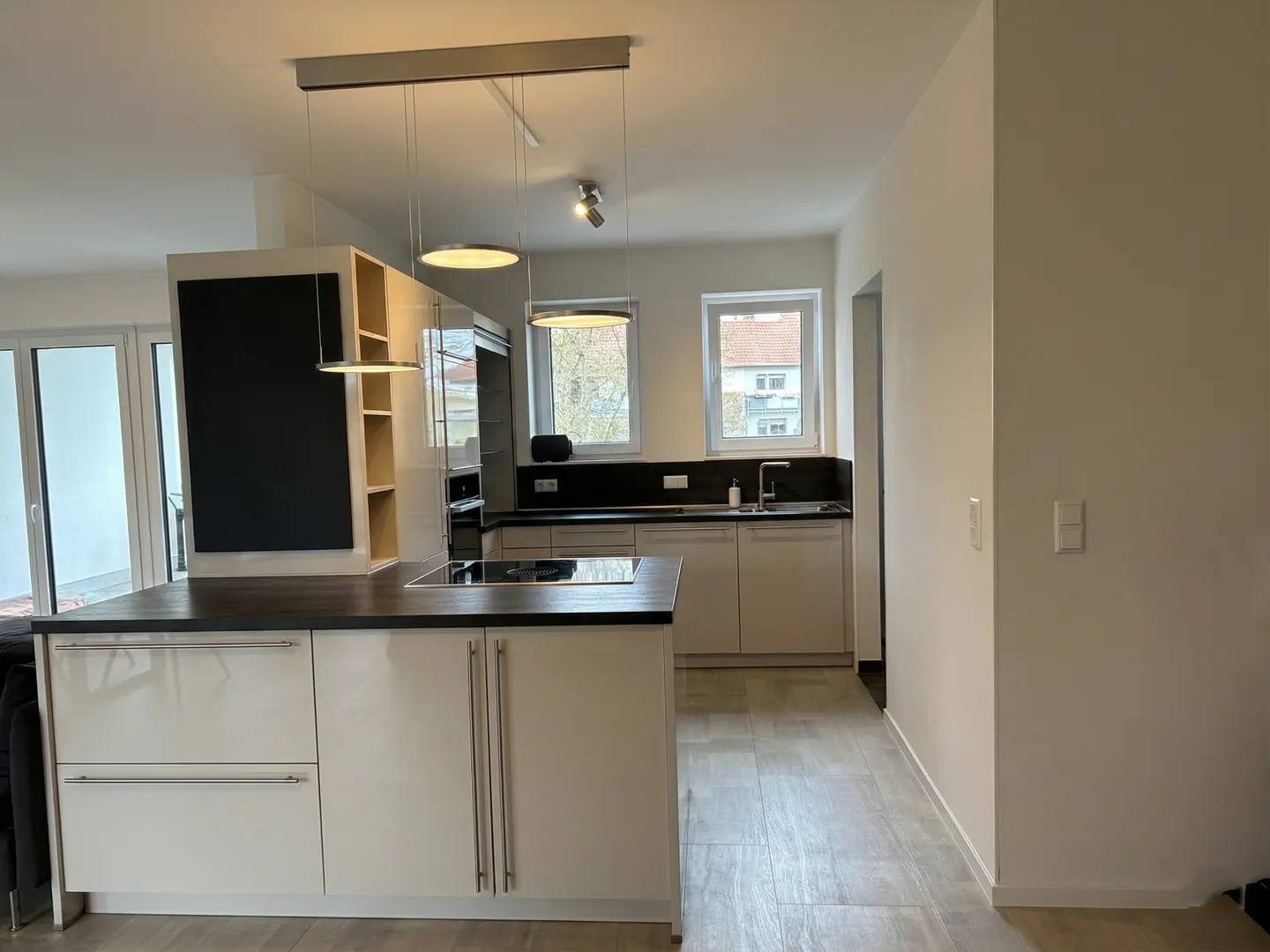 Bright, modern kitchen with an island, light wood cabinets, black countertops, and stainless steel appliances. Large windows provide natural light.