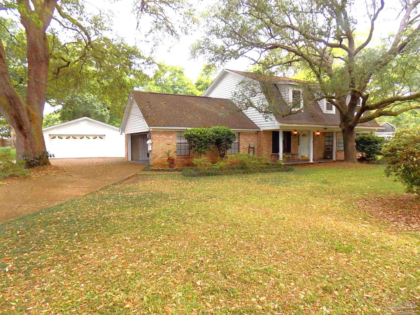 A brick house with a brown roof and white trim sits on a large lawn with trees. A white garage is visible in the background.