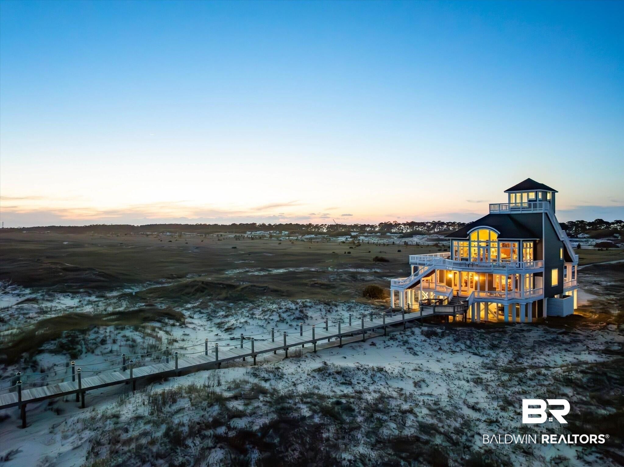 A multi-story coastal home with glowing interior lights sits on a sandy dune at dusk, connected by a long wooden walkway.