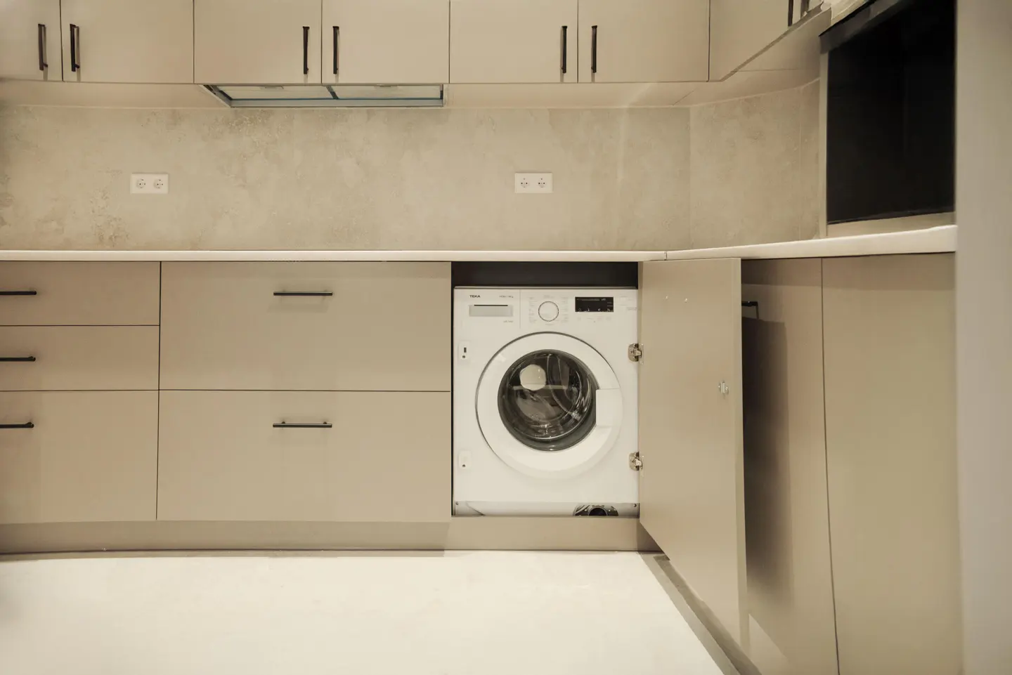 Modern kitchen with beige cabinets and black hardware. A white washing machine is built into the cabinetry.