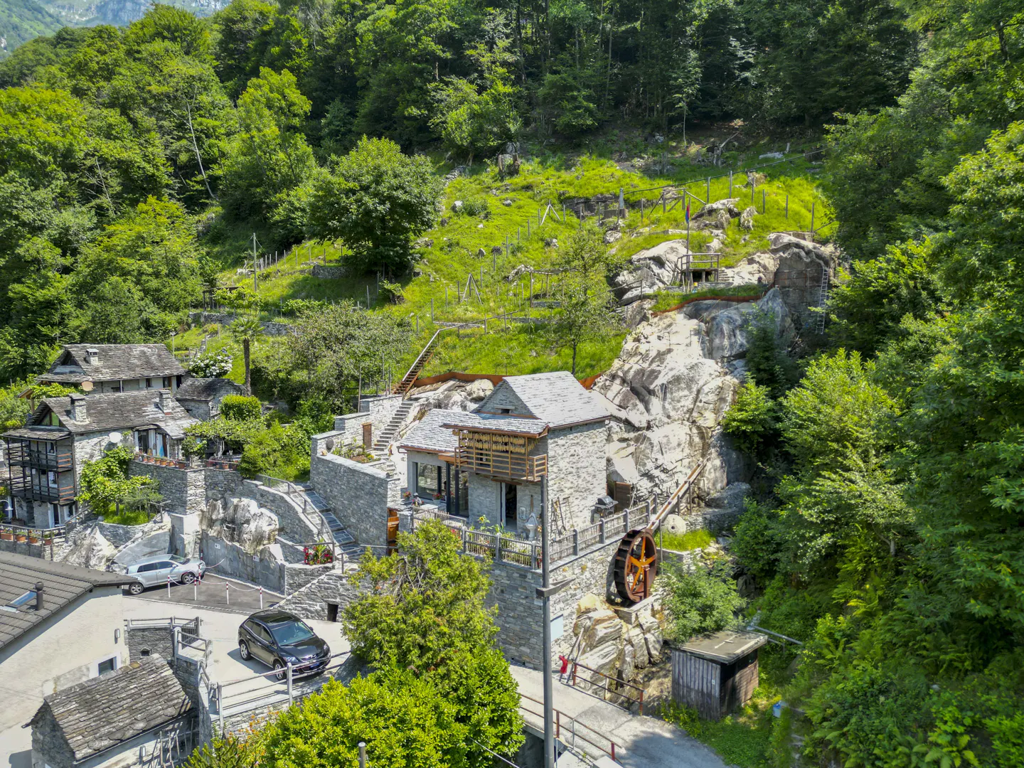 Aerial view of a stone house with a water wheel in a lush green forest setting.