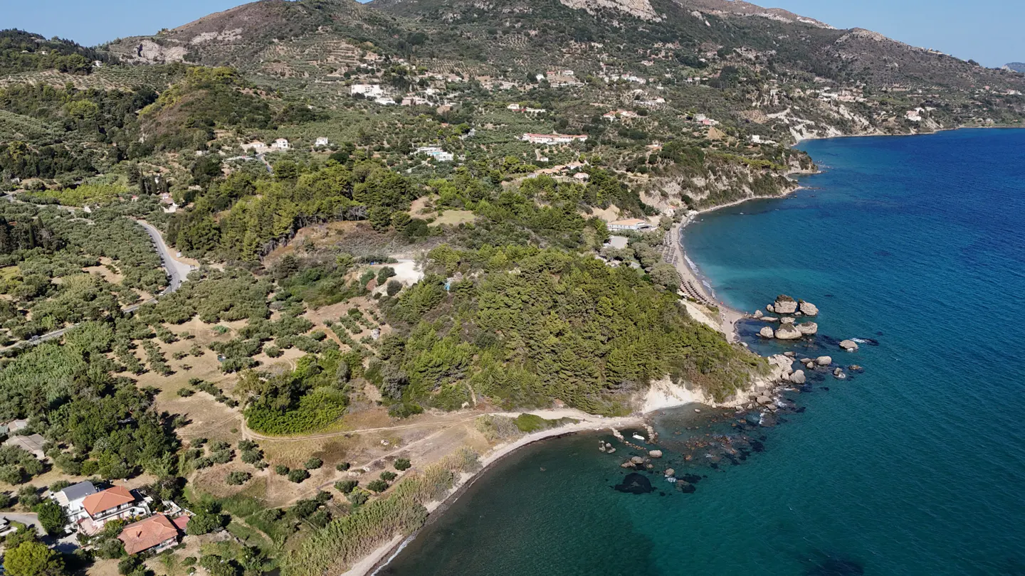 Aerial view of a green, hilly coastline meeting the turquoise sea, with houses nestled among the trees.