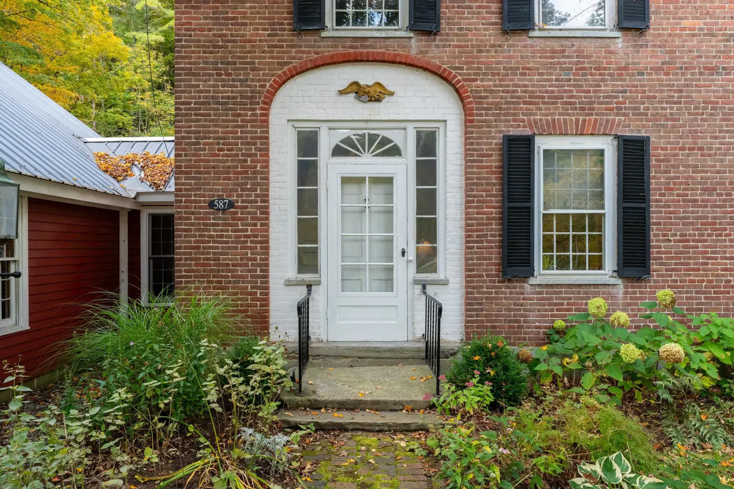 Brick house exterior with a white door framed by an arch and sidelights. Black shutters flank a window. Lush greenery in the front yard.