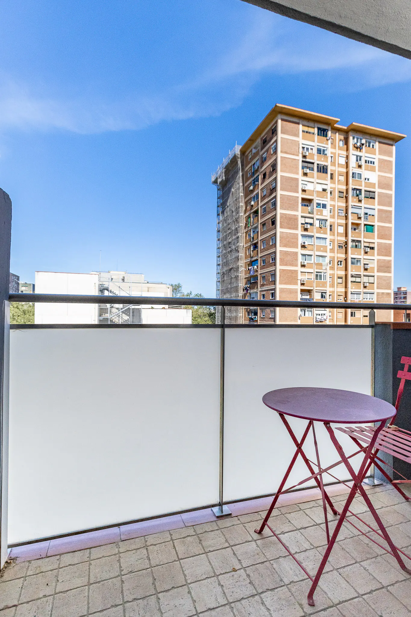 Balcony view with red table and chair, white frosted glass railing, and a tall brick building under a blue sky.