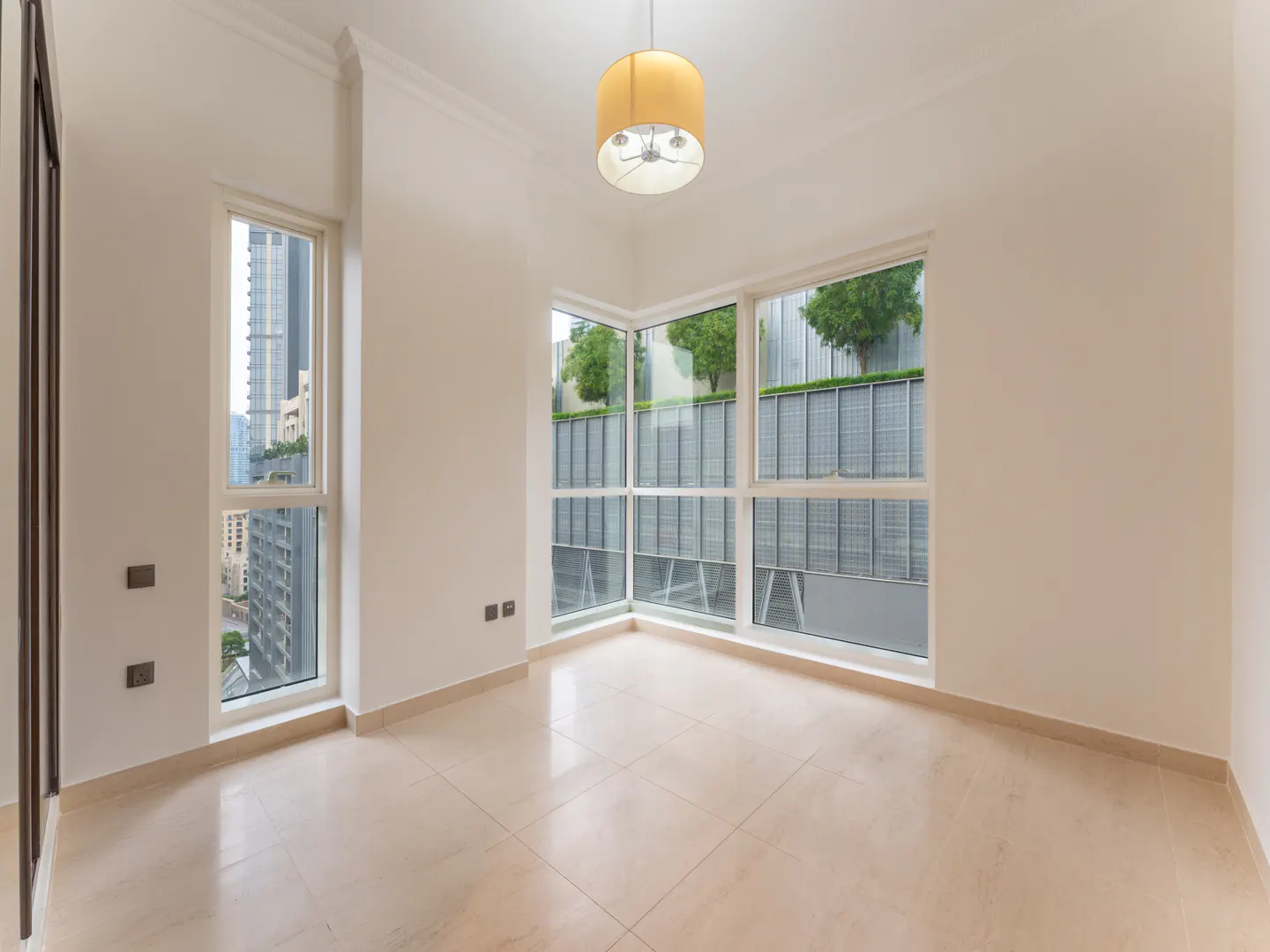 Bright, empty room with beige tile floor, white walls, and large windows showing city and greenery. A yellow drum pendant light hangs from the ceiling.