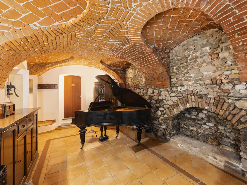 Interior view of a room with a brick vaulted ceiling, stone wall, and a black grand piano on a tiled floor.