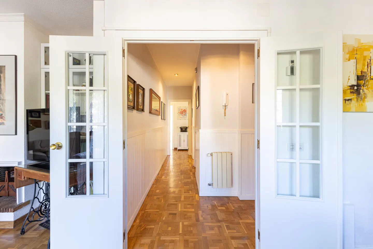 Open white French doors reveal a hallway with parquet floors and white wainscoting. Framed art lines the left wall.