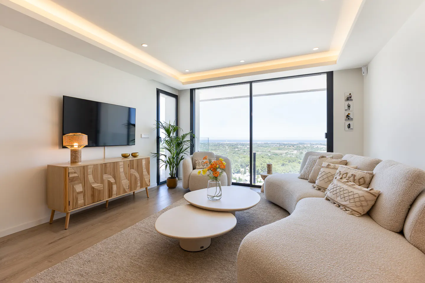 Bright living room with a curved white sofa, two round tables, and a wooden cabinet under a TV. Large windows overlook a green landscape.