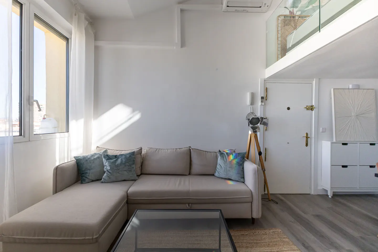 Bright living room with a beige sectional sofa, blue pillows, and a glass coffee table. A window and white walls add to the light airy feel.