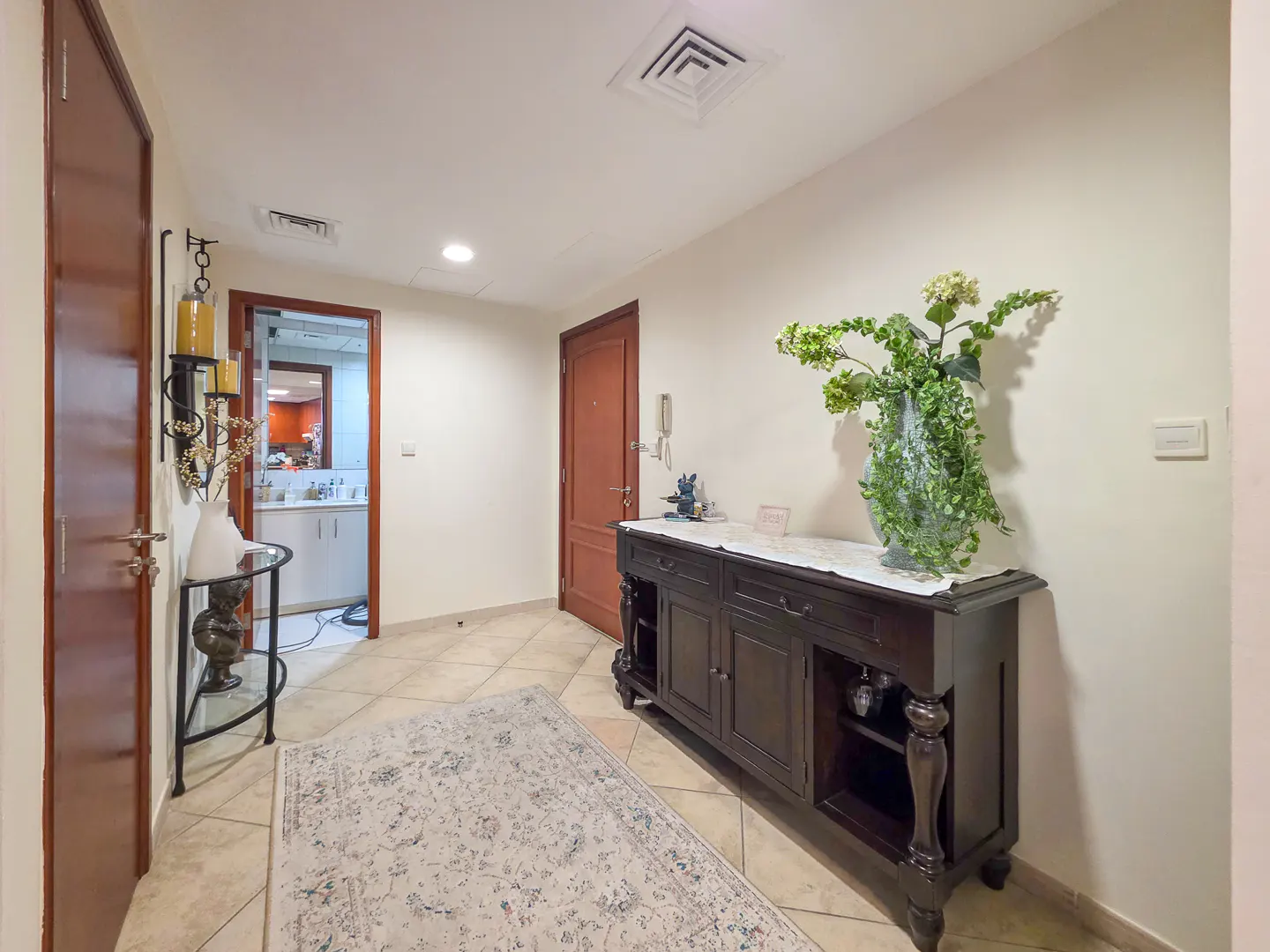 Hallway with beige tile floor, a dark wood console table with a large floral arrangement, and a patterned rug.