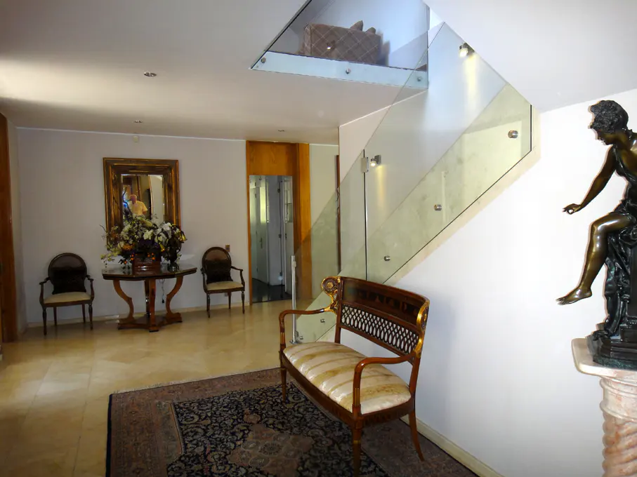 A bright foyer with a glass staircase, a bronze statue, and a wooden bench on a patterned rug.