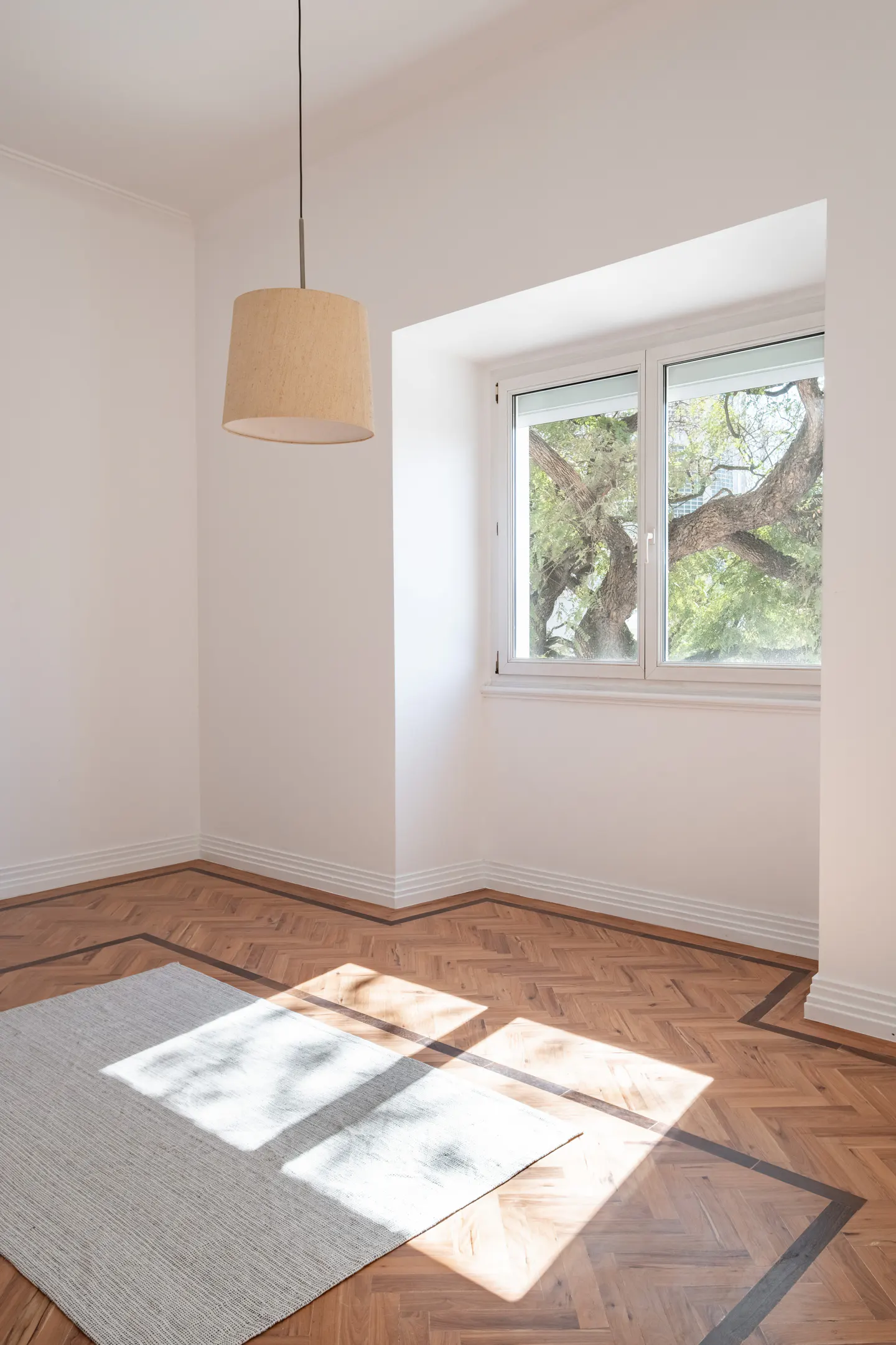 Bright, empty room with herringbone wood floors, a gray rug, and a tan pendant light. A window shows a large tree outside.