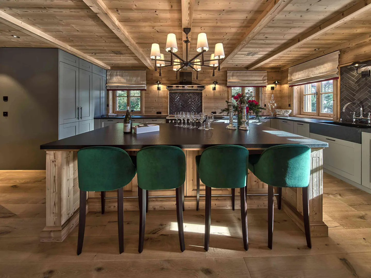 Rustic kitchen with wood-paneled ceiling, large island, four green velvet bar stools, and a modern chandelier.
