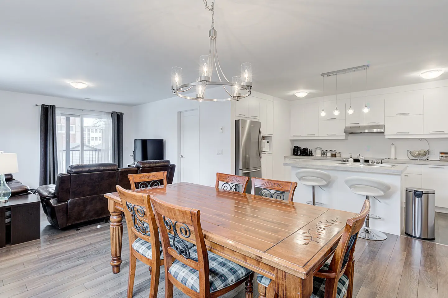 Bright, open-concept living space with a wooden dining table, plaid chairs, and a modern chandelier. White kitchen and brown leather sofa in background.