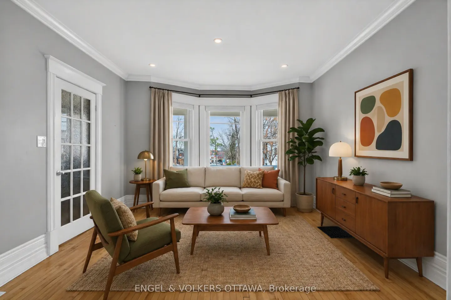 Living room with hardwood floors, gray walls, and a bay window. A cream sofa, wood furniture, and a green chair furnish the space.