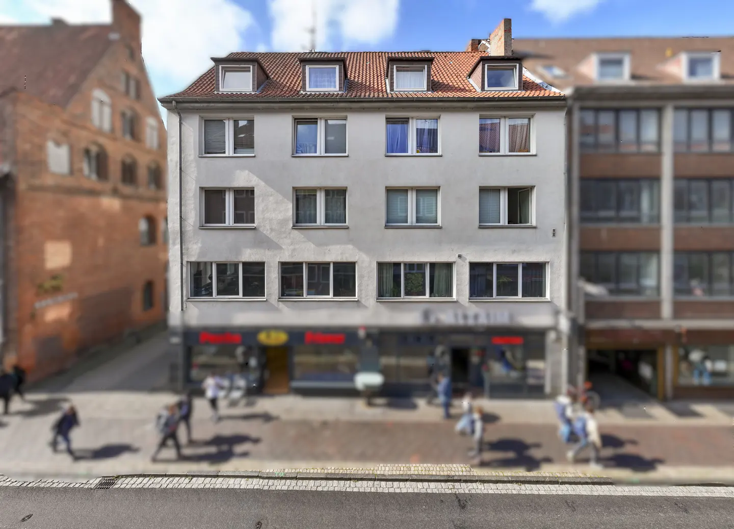 A four-story gray building with a red tile roof and dormer windows above a storefront on a busy city street.
